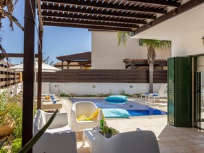 Un área de piscina con sillones reclinables, una pérgola y vistas a un edificio al fondo.