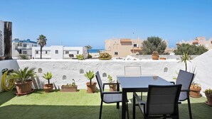 A terrace with a table and chairs, potted plants, and a view of buildings and the sea.