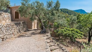 A stone building with a gravel path leading to a wooden door, surrounded by trees and a mountainous backdrop.