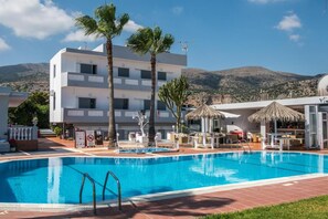 Una piscina con agua cristalina y azul, rodeada por un área de descanso con tumbonas y sombrillas, y un edificio con varios pisos y balcones al fondo.