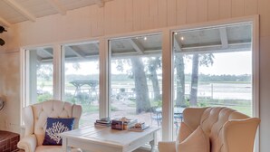 Una habitación con dos sillones blancos, una mesa blanca con libros, y ventanas amplias que dan a un área verde.