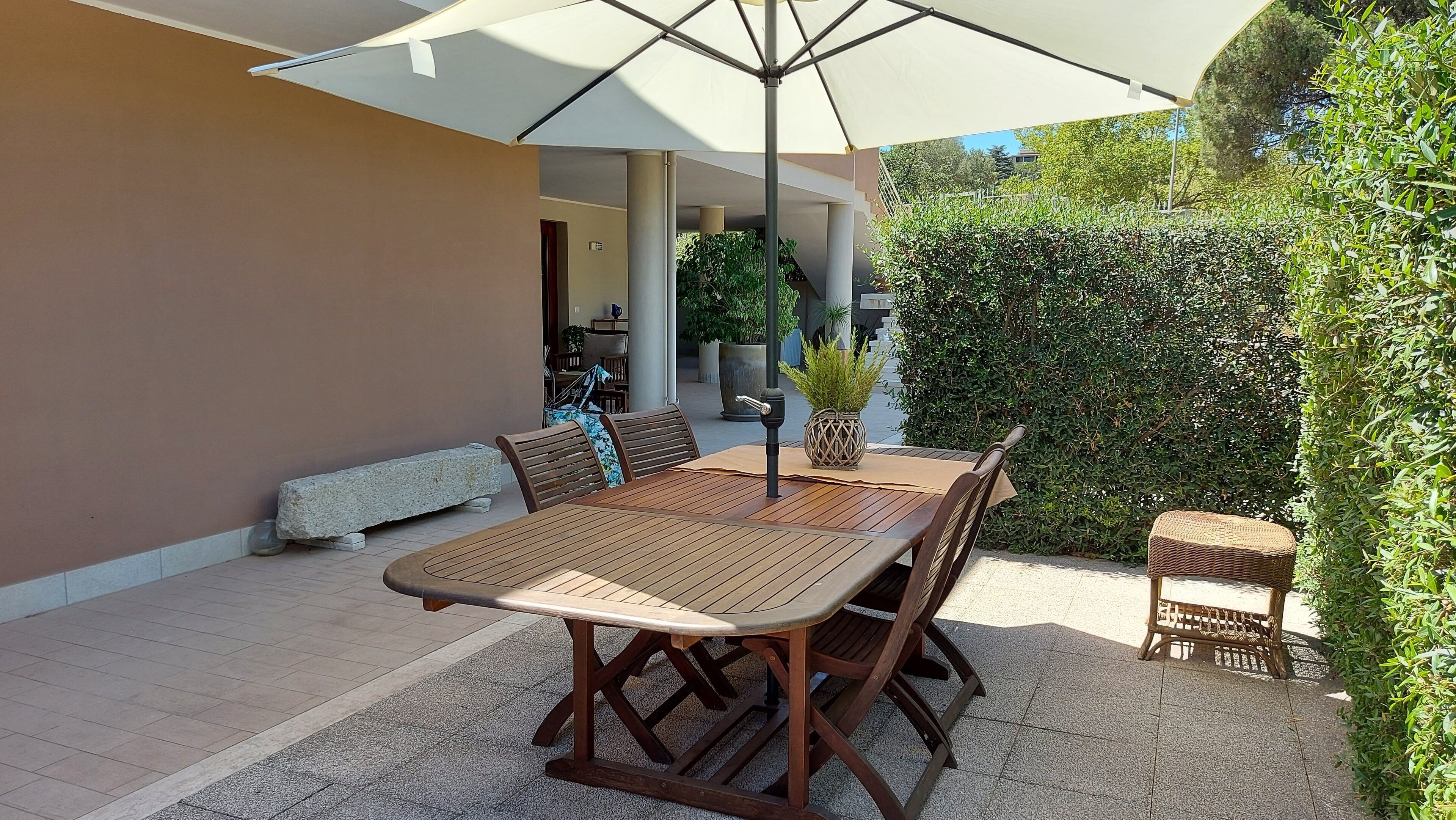 An outdoor patio with a wooden table and chairs, a large umbrella, and a stone bench.