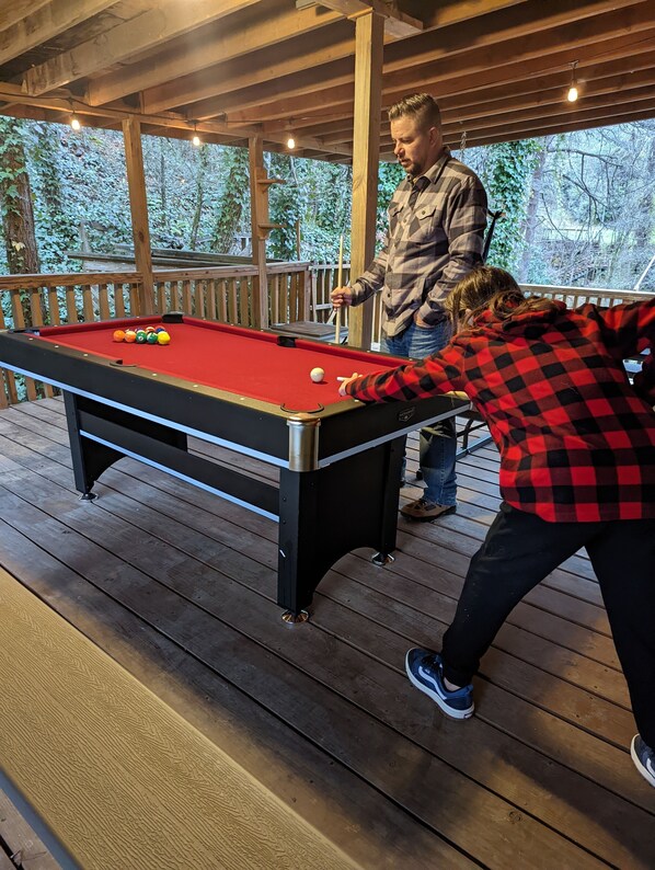 A pool table on a wooden deck with a railing and string lights overhead. - Stay in the Treehouse Lodge! Hot tub, game room, fire place, wrap around porch! (Bryson City)
