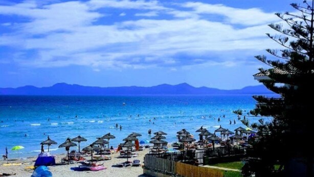 Una playa con sombrillas y sillas reclinables, una valla y vista al mar y montañas distantes.