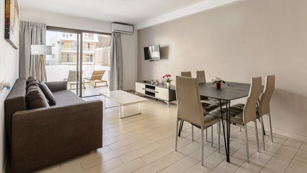 A modern living room with a sofa, dining table, chairs, a flat-screen TV, and a balcony with a view of buildings.