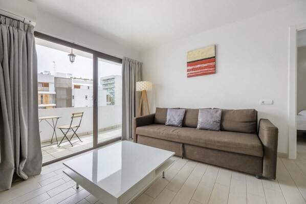 Un salon moderne avec un canapé, une table basse et une vue sur un balcon extérieur, accessible par une grande porte-fenêtre coulissante.