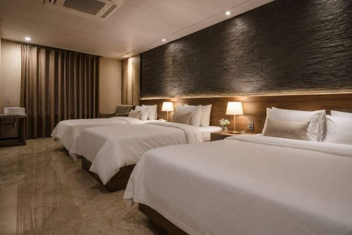 A hotel room with three beds, a wooden headboard, a nightstand with a lamp, and a large window with curtains.