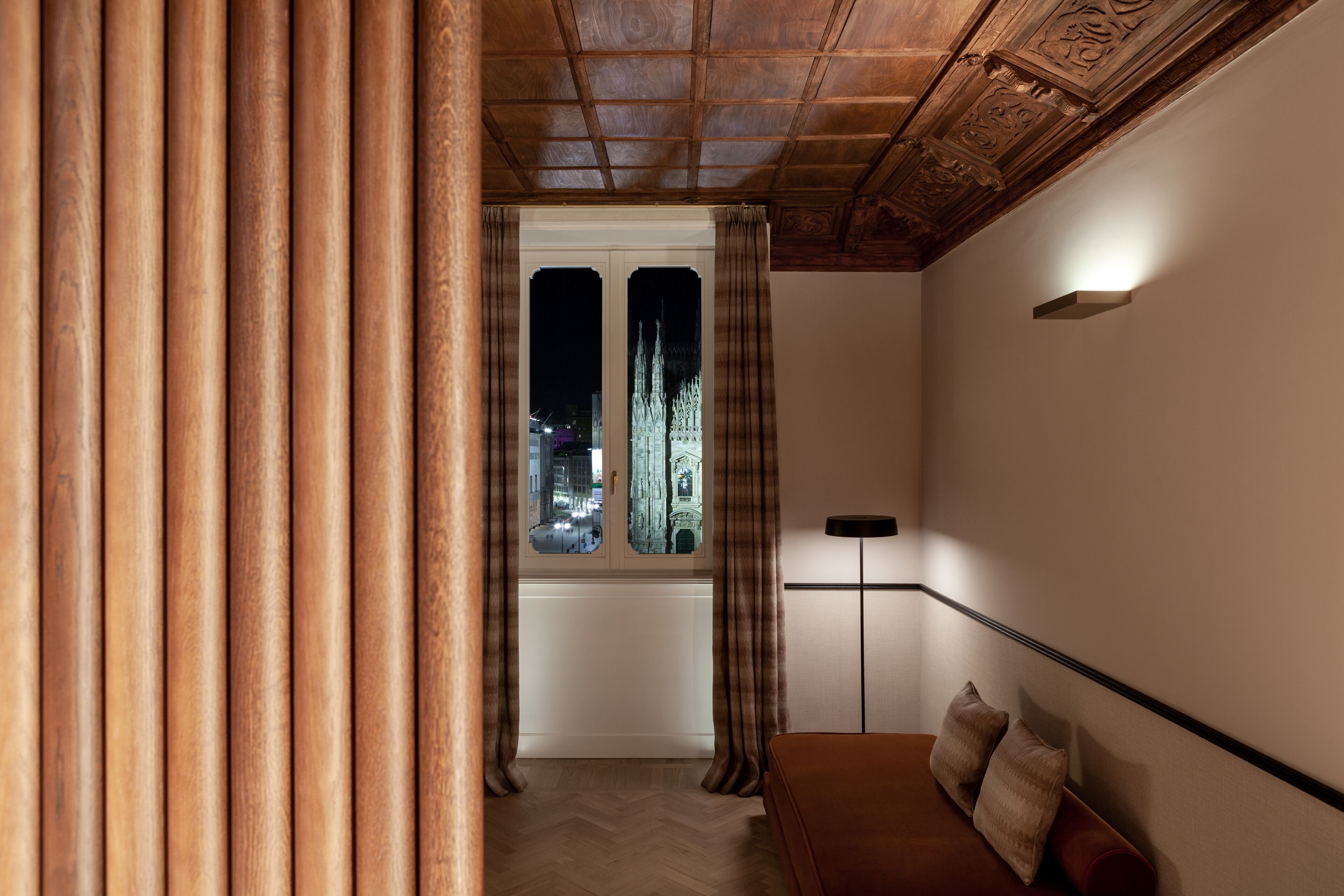 A room with a wooden ceiling, a window with curtains, a sofa with cushions, and a floor lamp.