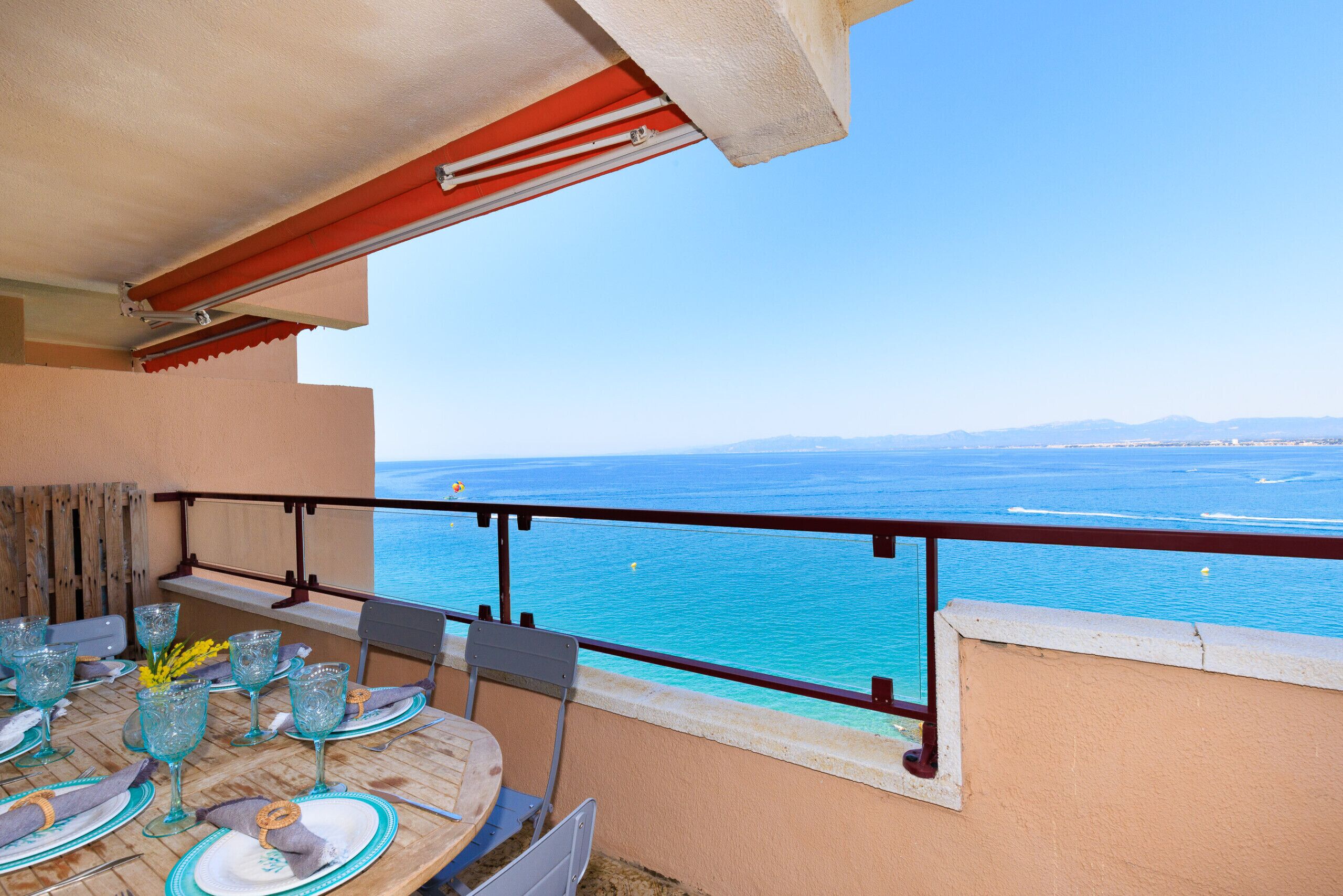 A balcony with a table set for dining, overlooking a clear blue sea and distant mountains.