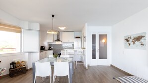 Moderni keittiö, jossa on valkoiset kaapit, keskellä oleva keittiösaareke ja ruokailutila valkoisine tuoleineen.