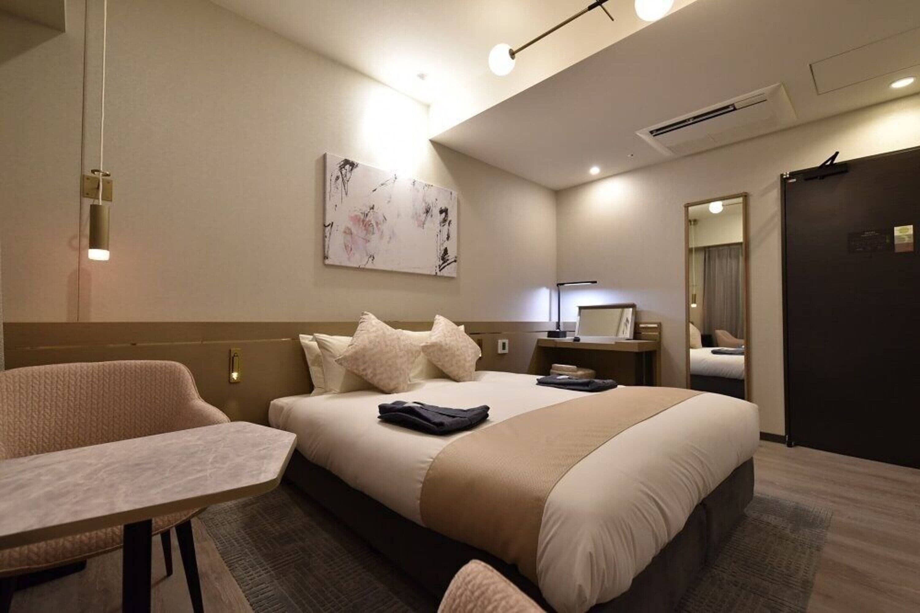 A modern hotel room with a large bed, a desk, and a chair. There is a painting on the wall, a desk lamp, and a mirror.