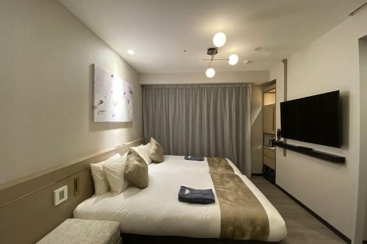 A hotel room with a bed, a flat-screen TV mounted on the wall, a ceiling light fixture, and a window with curtains.