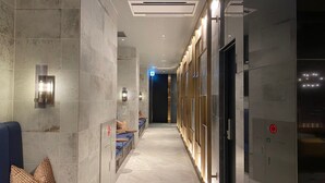 A modern hotel corridor with a bench, wall-mounted light fixtures, and a door with a red emergency exit sign.