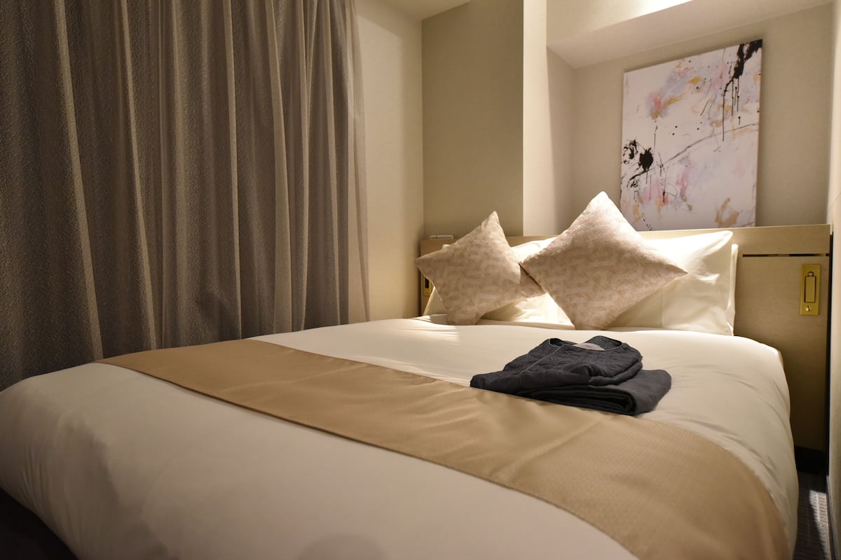 Een netjes opgemaakt bed met een beige sprei en twee kussens, een donkergekleurd kledingstuk gevouwen op het bed, en een groot abstract schilderij aan de muur.