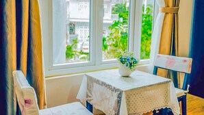 A room with a table and chair, a window with curtains, and a view of buildings and greenery outside. - Jolie House II - Villa 2 Double Rooms - Charming (Thành phố Đà Lạt)