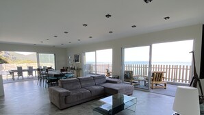 Un vaste espace de vie comprenant un grand canapé d’angle gris, une table basse en verre et un lampadaire blanc. La pièce est dotée d’une grande porte-fenêtre coulissante qui donne sur un balcon offrant une vue sur la mer.
