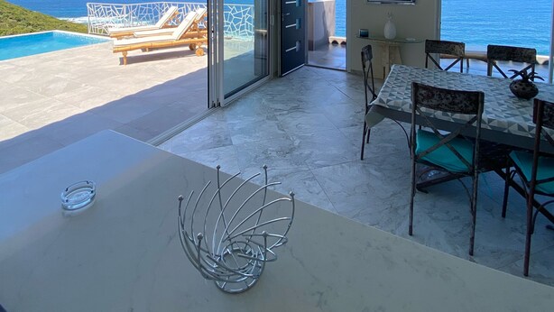Une pièce spacieuse dotée d’une grande porte-fenêtre coulissante donnant sur un balcon avec vue sur l’océan. On y trouve un coin repas avec une table et des chaises, ainsi qu’une sculpture métallique décorative posée sur la table.