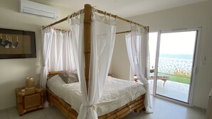Une chambre dotée d’un lit à baldaquin, d’une grande fenêtre donnant sur la mer et d’une petite table de chevet en bois avec une lampe.