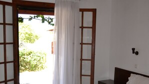 Un dormitorio con una cama grande, una mesita de noche de madera y una silla. Se ve una ventana con cortinas y vistas al exterior con vegetación.