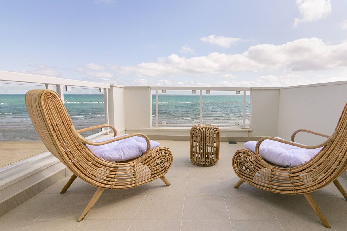 A balcony with two wicker chairs and a small table, overlooking the ocean.