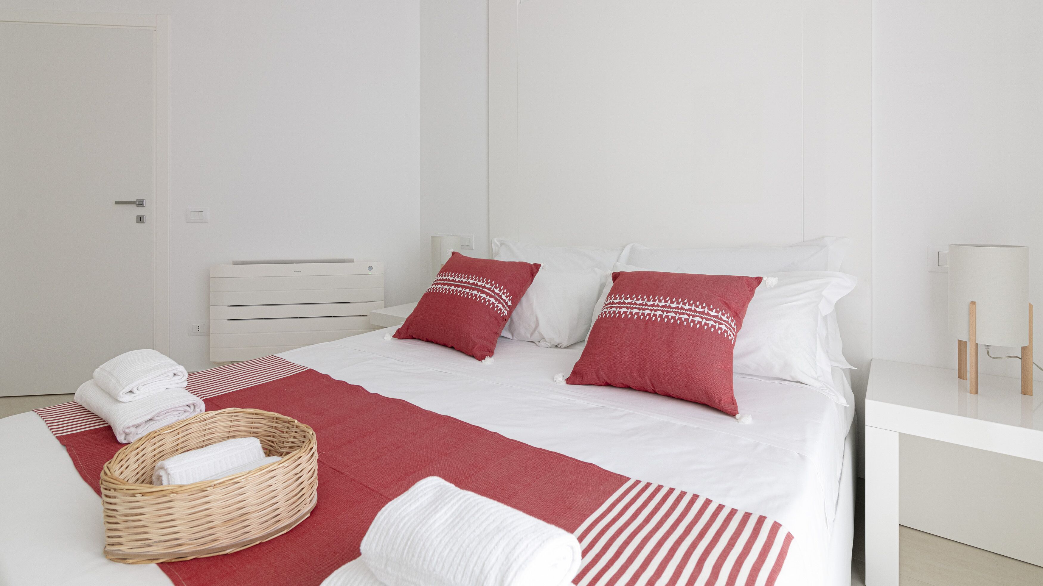 Une chambre d’hôtel avec un lit, du linge de lit blanc, des oreillers rouges et blancs, un panier en osier contenant des serviettes et une table de chevet avec une lampe.