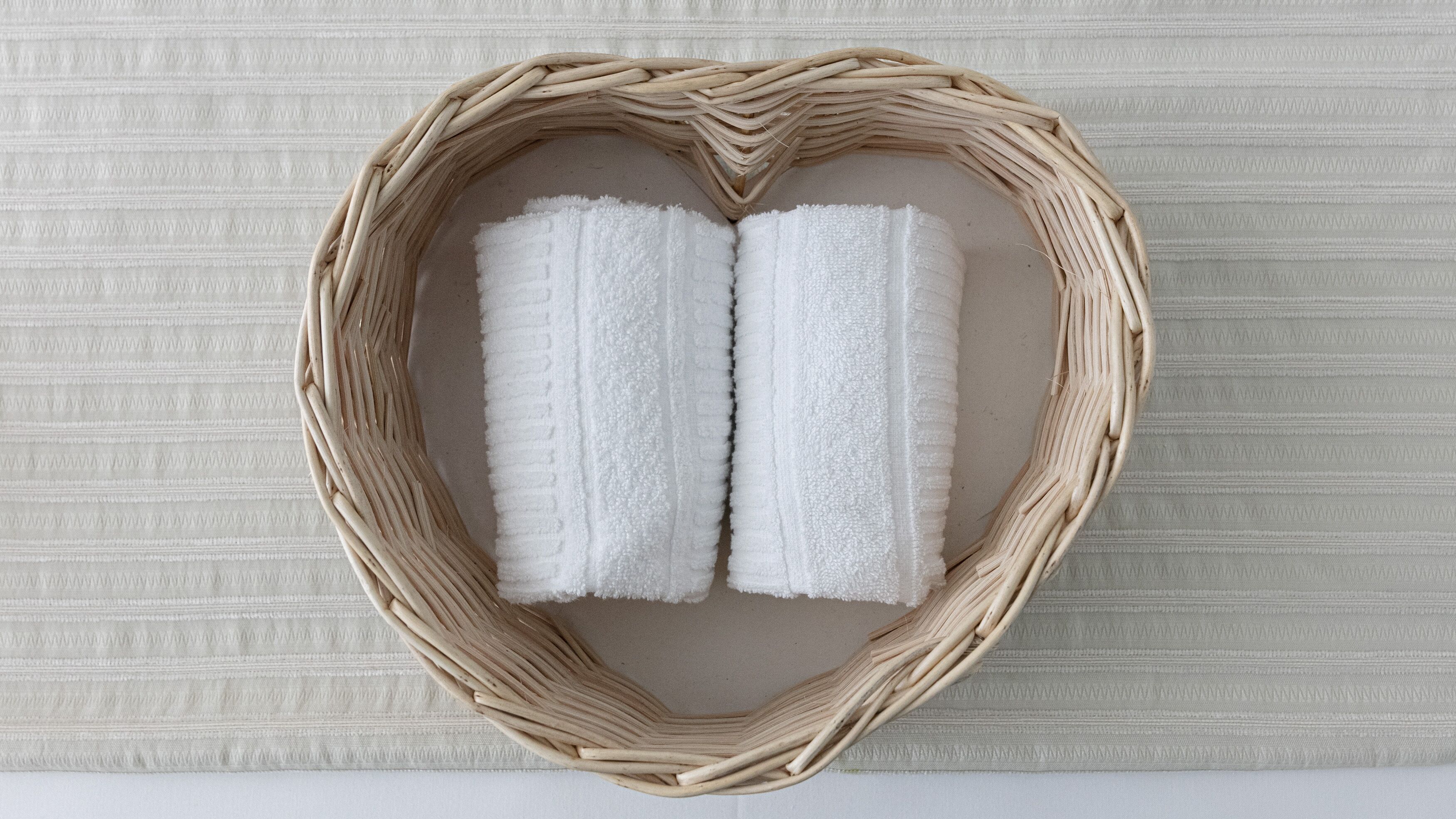 Una cesta de mimbre con forma de corazón que contiene dos toallas blancas frente a una pared texturizada.
