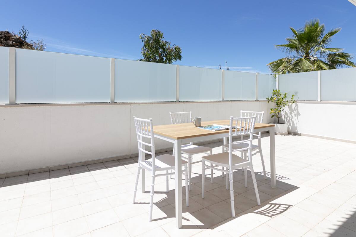 A patio with a wooden table and white chairs, a white wall, and a palm tree in the background.