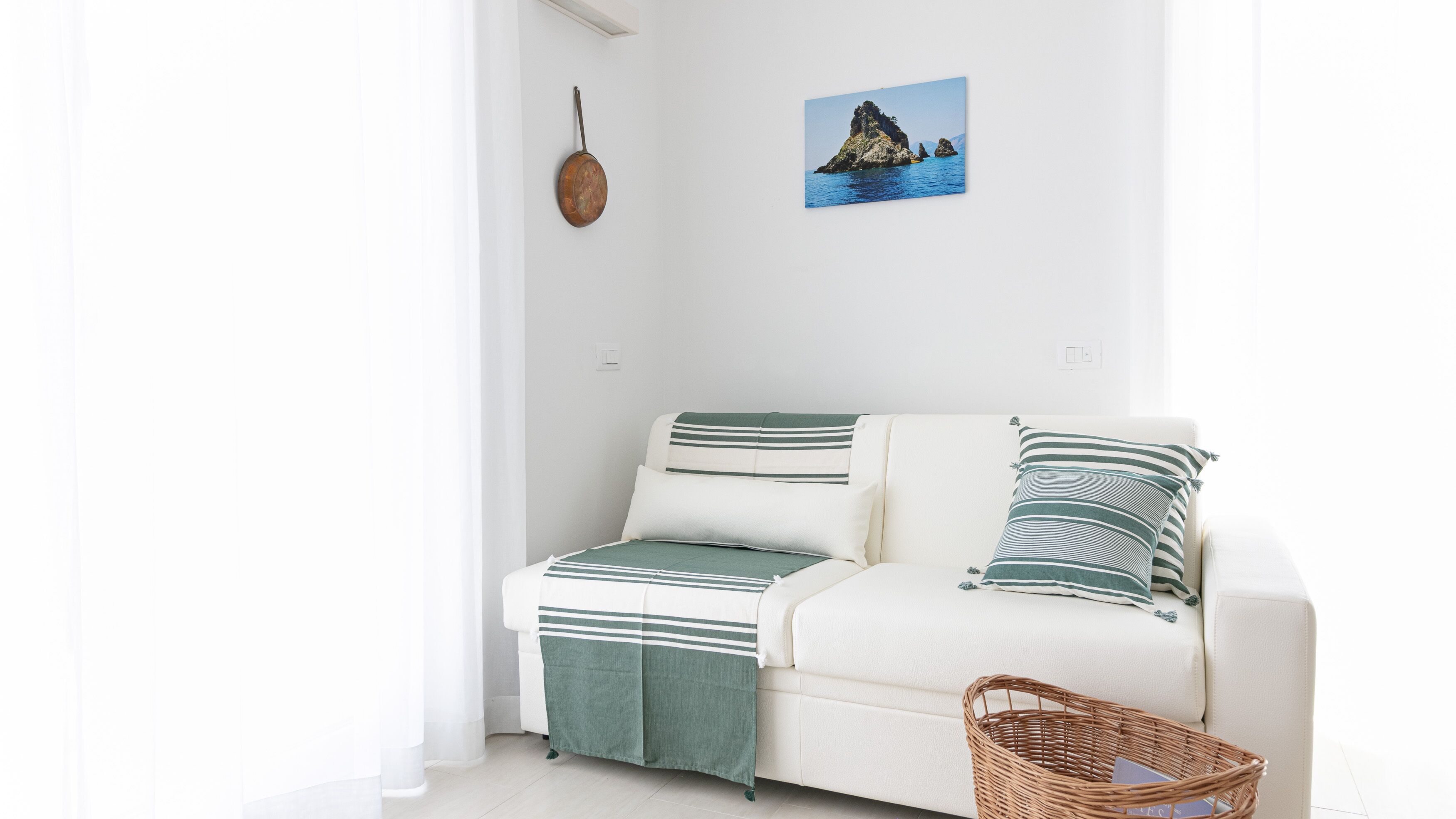 A white sofa with striped pillows and a green throw blanket, a wicker basket, and a framed picture of a coastal landscape on the wall.