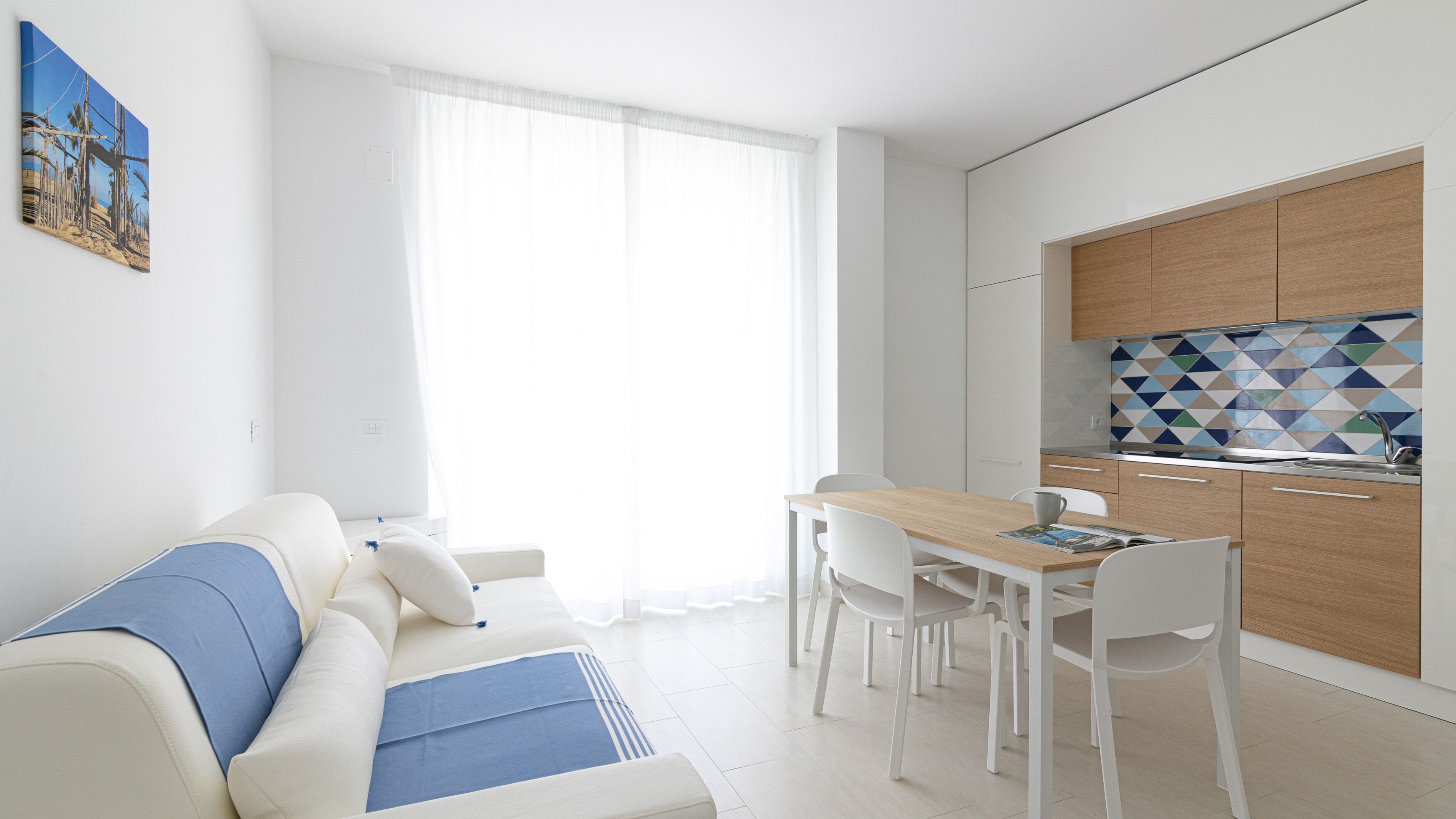 Un salon moderne comprenant un canapé, une table à manger avec des chaises, un coin cuisine avec un mur recouvert de carreaux, et une grande fenêtre agrémentée de rideaux légers.