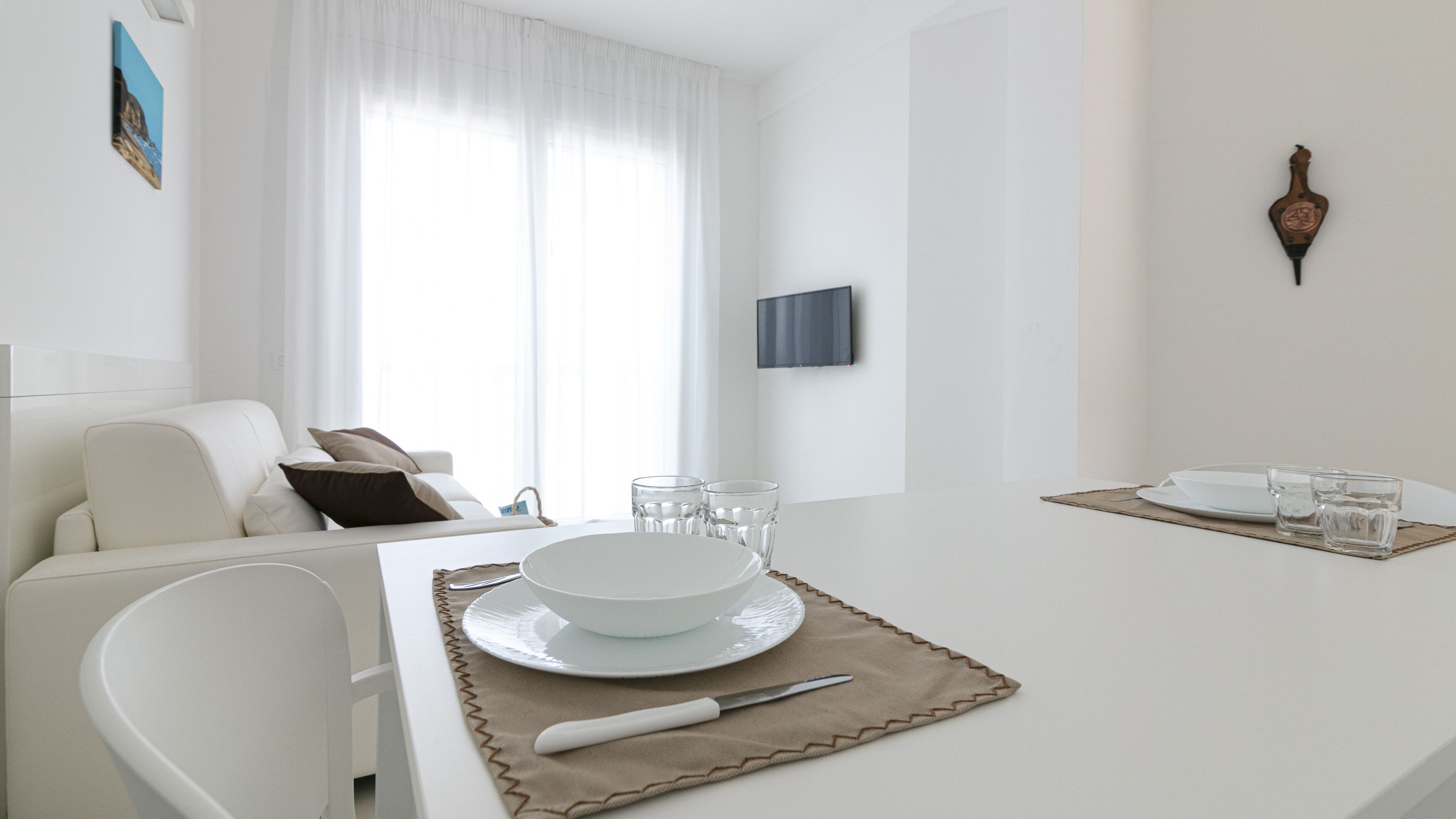 Un comedor moderno y minimalista con una mesa blanca dispuesta para dos personas, incluyendo platos, vasos y cubiertos.