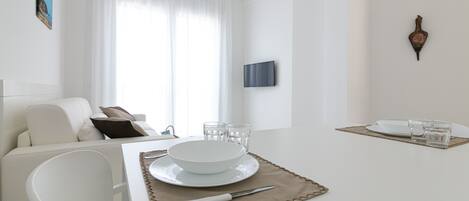 Un comedor moderno y minimalista con una mesa blanca dispuesta para dos personas, incluyendo platos, vasos y cubiertos.
