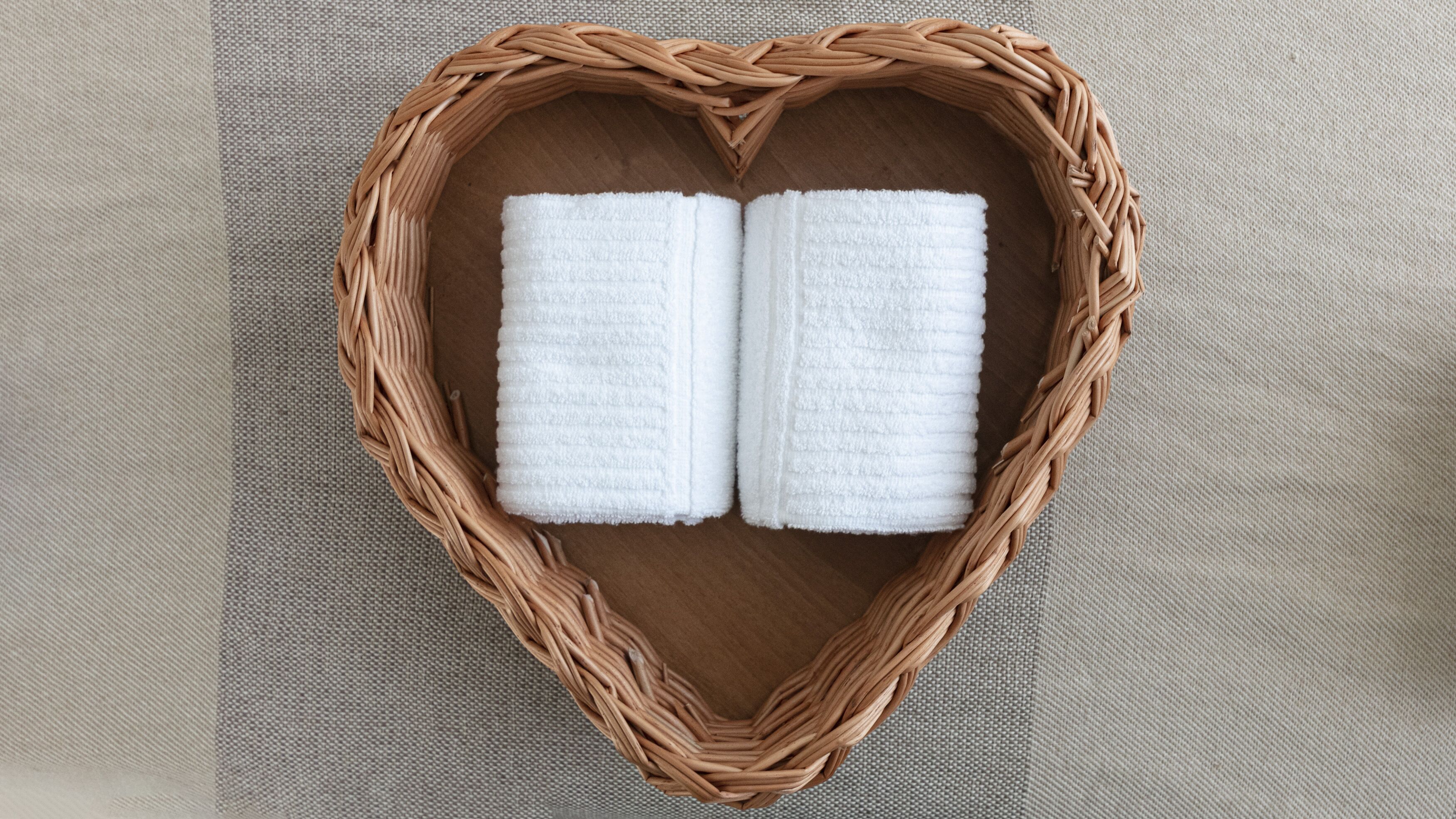 Una cesta de mimbre con forma de corazón que contiene dos toallas blancas sobre una superficie texturizada.