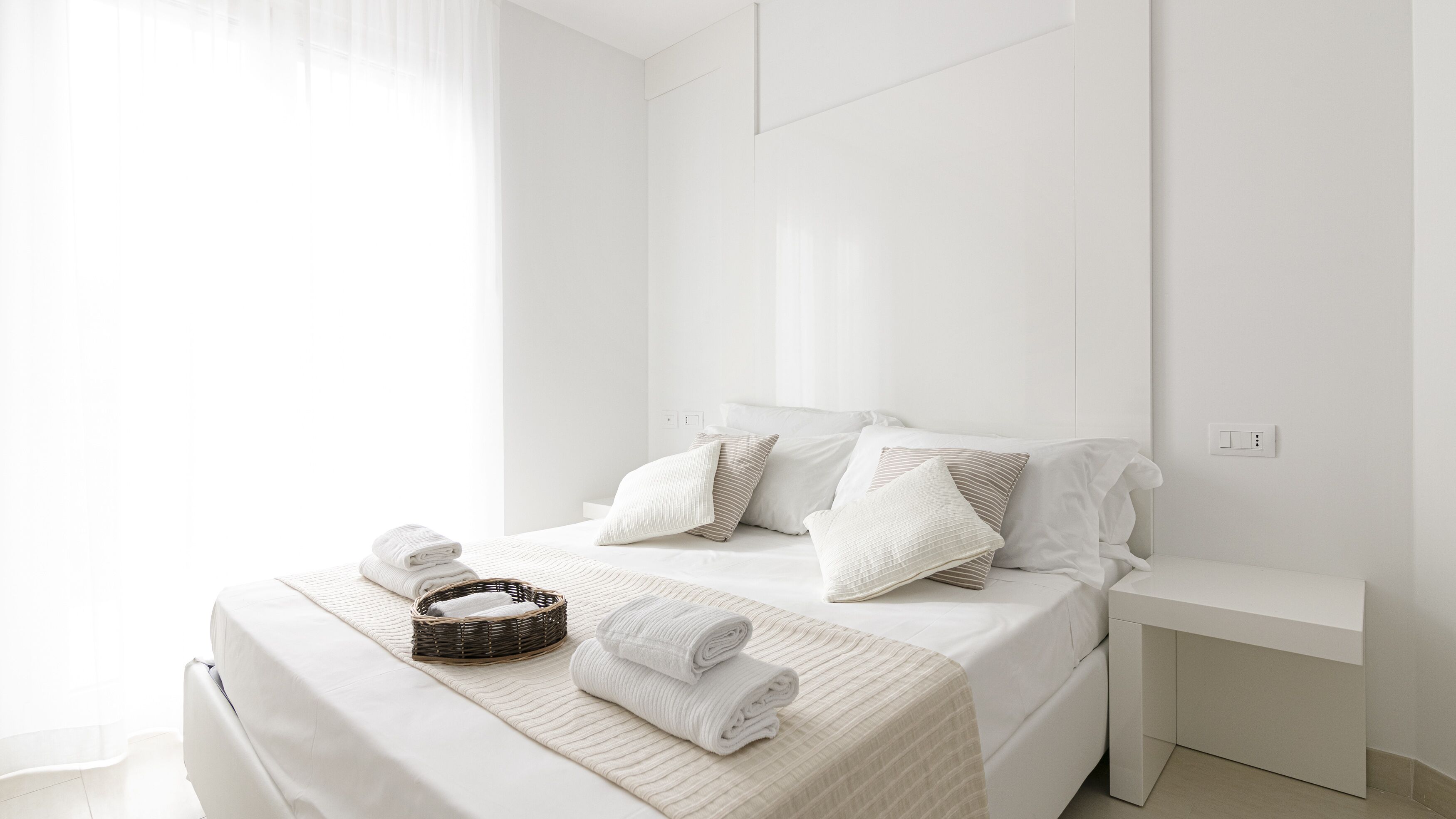 Une chambre à coucher minimaliste, aux murs blancs, avec un lit soigneusement fait, une table de chevet et un panier en osier contenant des serviettes.