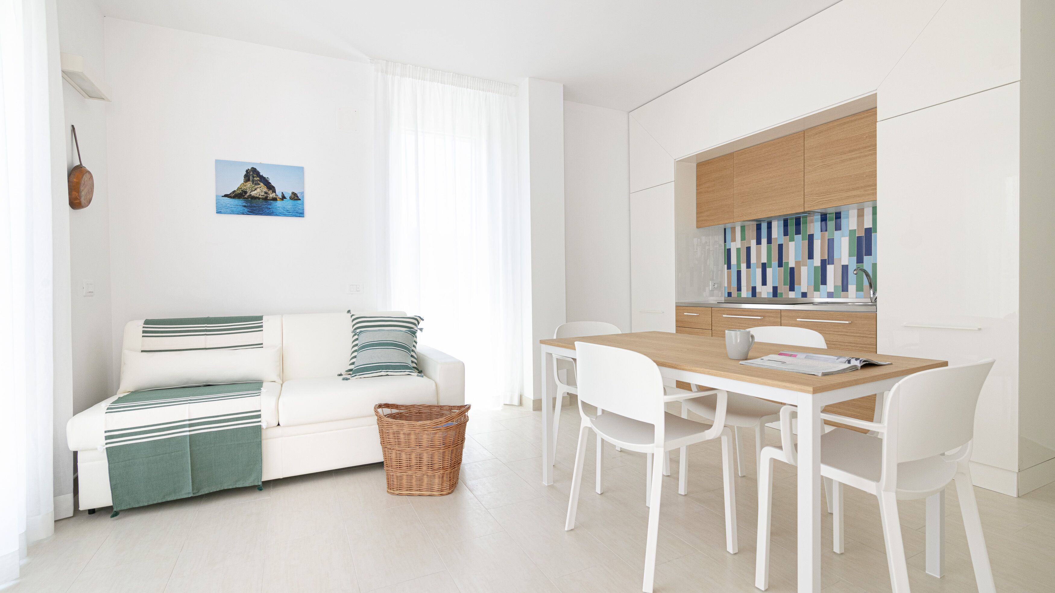 A modern, minimalist living space with a white sofa, a dining table with chairs, and a kitchen area with wooden cabinets and a colorful backsplash.