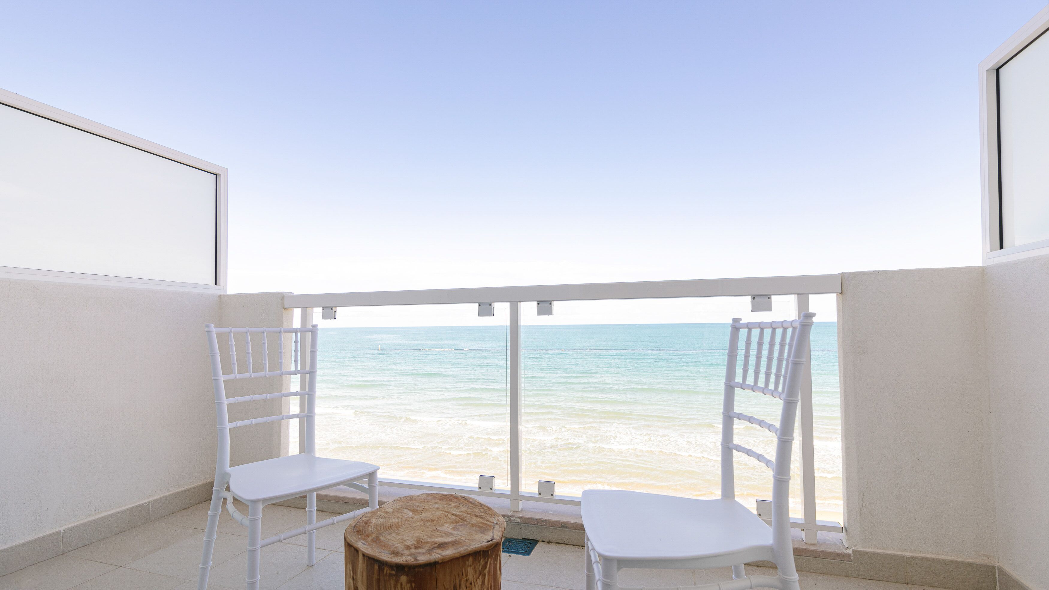 Un balcón con dos sillas blancas y una pequeña mesa de madera, con vistas a una playa y aguas tranquilas.