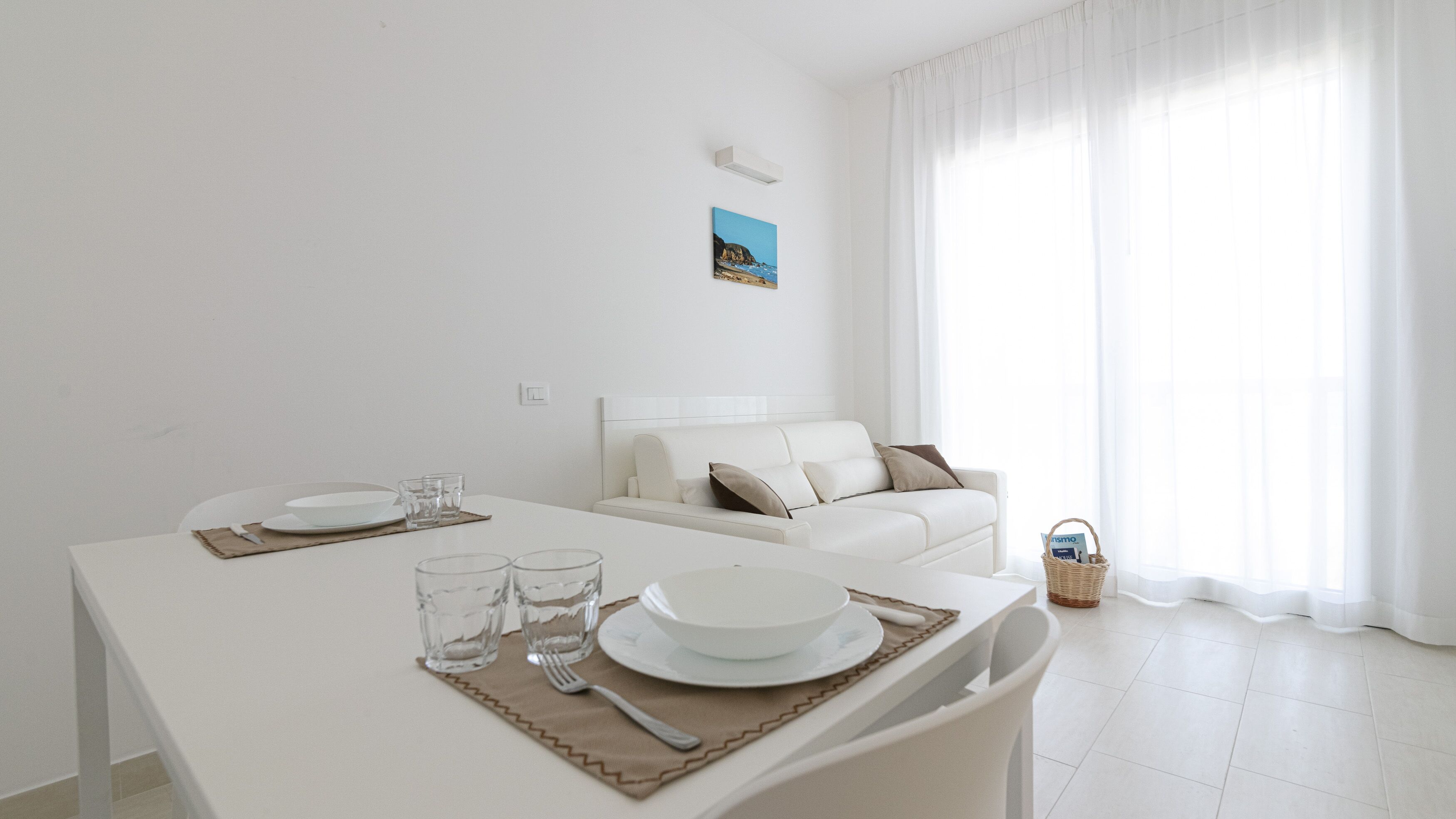 A modern, minimalist dining area with a white table set for two, a white sofa, and a large window with sheer curtains.