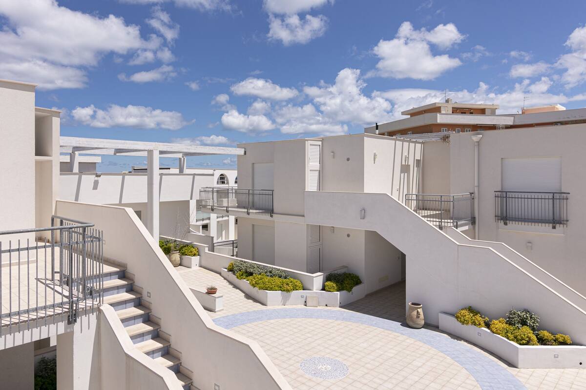 A modern residential building with multiple balconies, a central courtyard with potted plants, and a clear blue sky.
