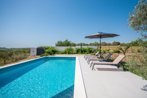A swimming pool with lounge chairs and an umbrella, surrounded by greenery and a clear sky. - Modern villa BlackWood in Crno, secluded area, near Zadar center, near the beach, private pool, en suite bathrooms (Crno)