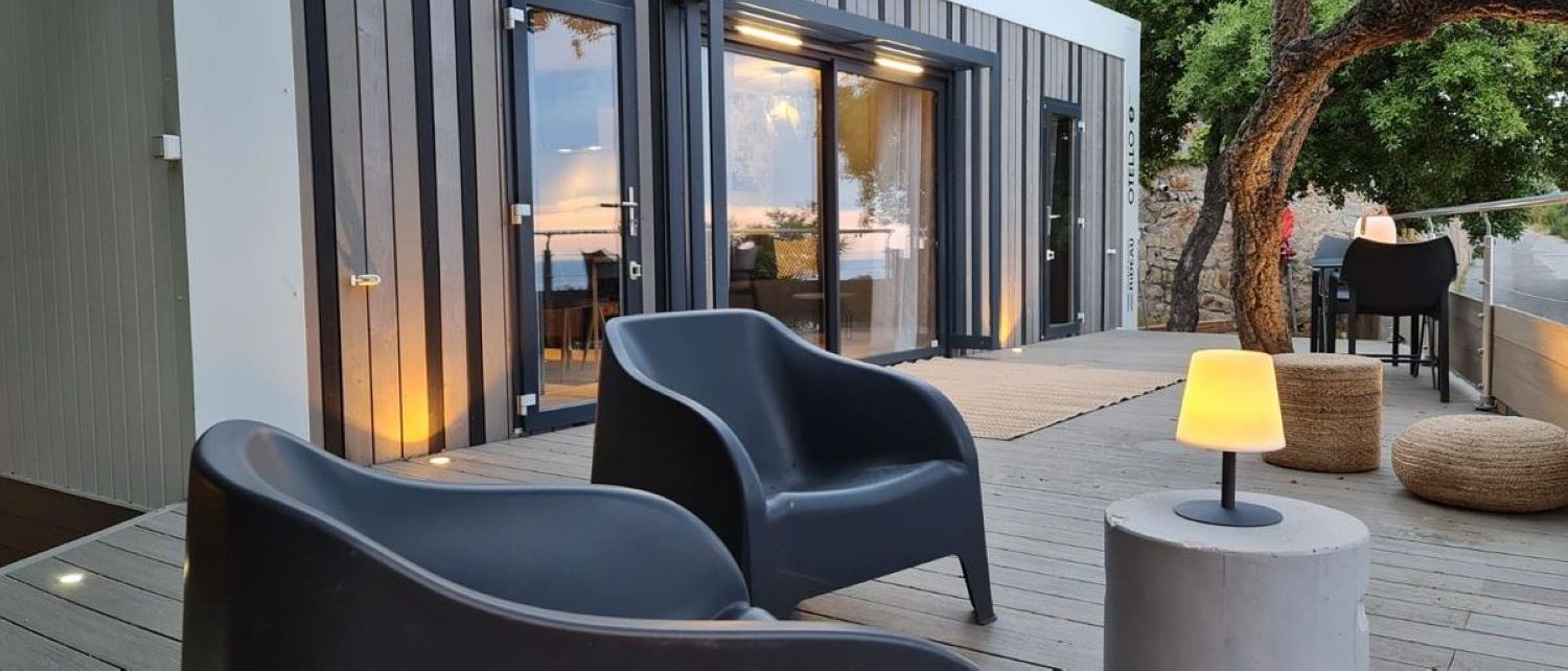 Ein moderner Sitzbereich im Freien mit zwei Stühlen, einem Tisch, einer Lampe und Blick auf ein Gebäude mit großen Glasfenstern.