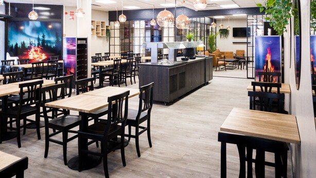 A modern restaurant interior with wooden tables and black chairs, a central bar area, and pendant lighting. - Hotelli Loimu (Raisio)
