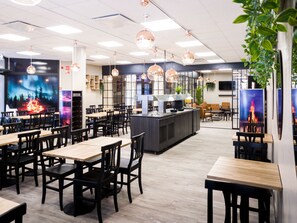 Un interior de restaurante moderno con mesas de madera y sillas negras, una zona de bar central e iluminación colgante.