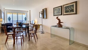Un coin repas avec une table dressée pour quatre personnes, entourée de chaises en bois. On y trouve également une table basse en verre sur laquelle est posée une sculpture. La pièce est dotée d’une grande fenêtre offrant une vue sur la mer.