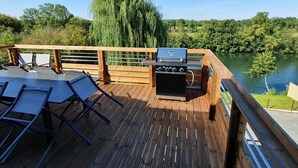 Een houten terras met een barbecue en twee loungestoelen met uitzicht op een meer.