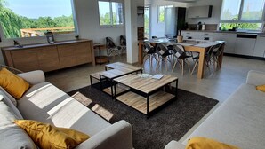 Een moderne woonkamer met een bank, salontafel en eethoek. Er is een keuken met een spoelbak en kasten. Grote ramen zorgen voor natuurlijk licht en uitzicht op de omgeving.