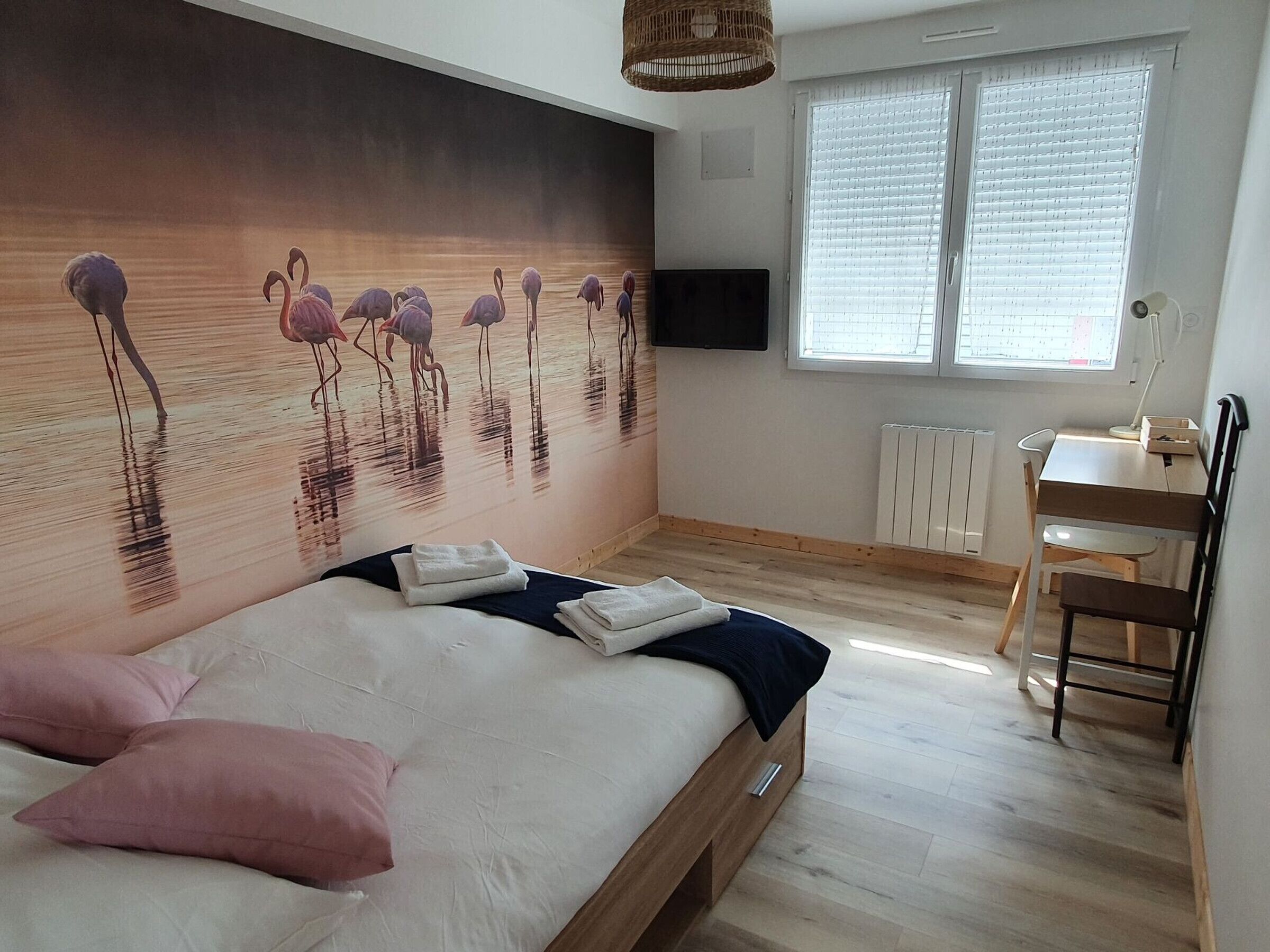 A bedroom with a large mural of flamingos, a bed with pillows, a desk, and a chair.