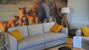 Een woonkamer met een grijze bank, gele kussens, een ventilator, een lamp en een groot schilderij van leeuwen.