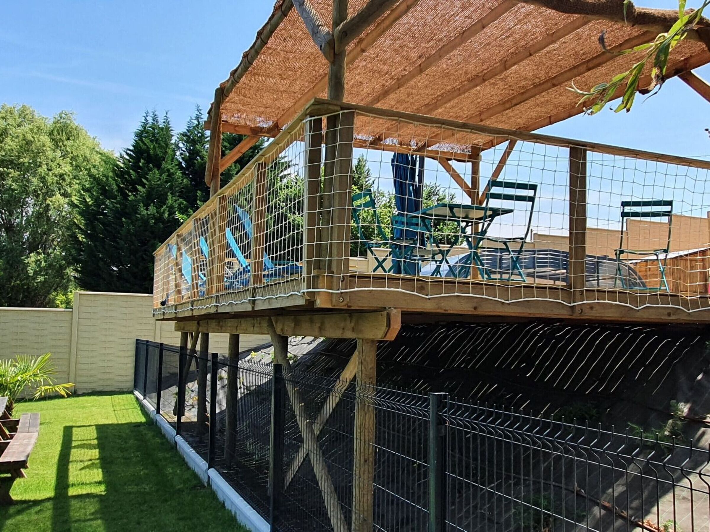Eine Holzterrasse mit einem Reetdach, umgeben von einem Metallzaun, sowie ein Tisch mit Stühlen.
