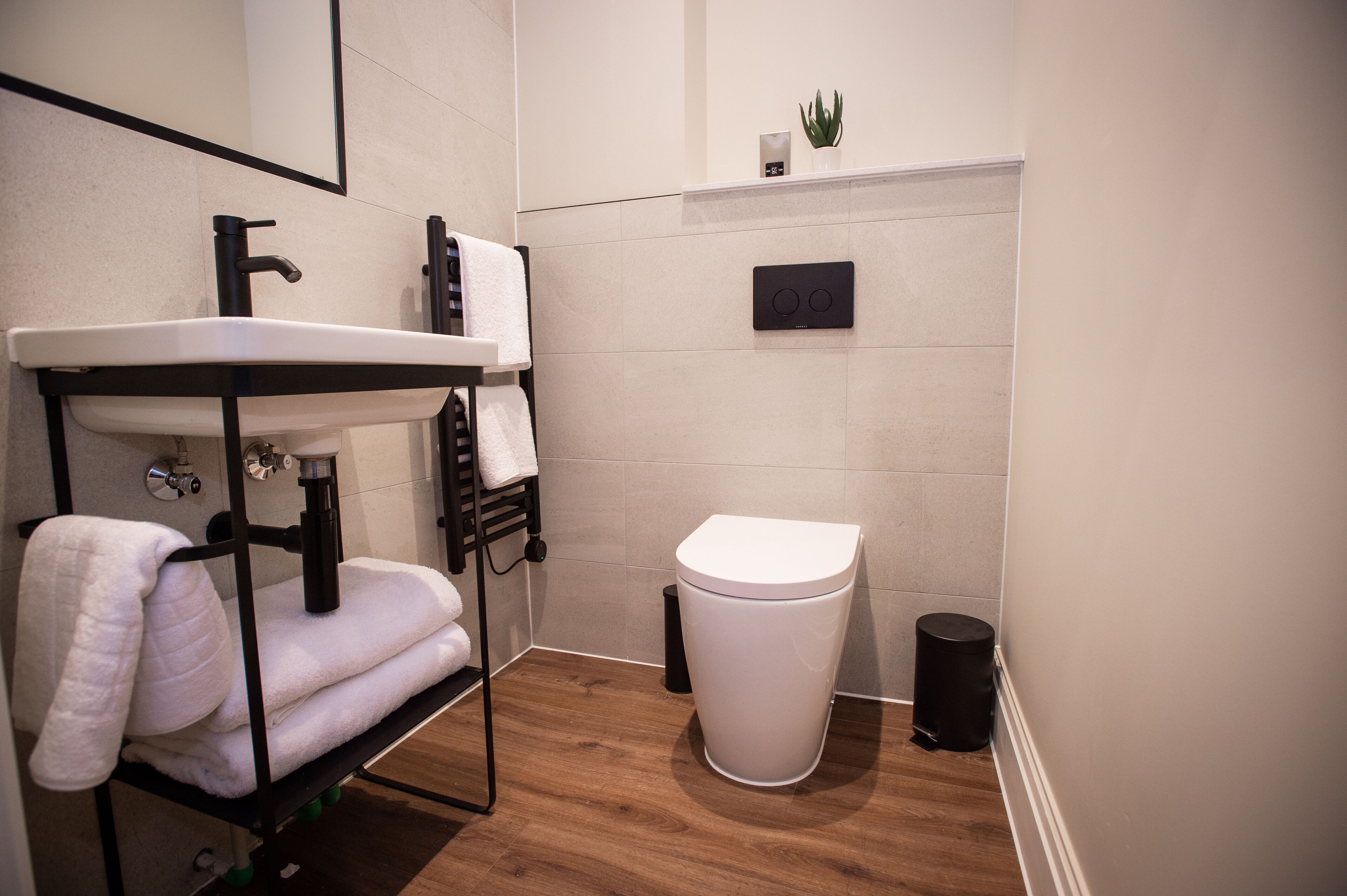 A compact bathroom with a white sink, black faucet, and a white toilet. There is a towel rack with folded towels, a small potted plant on the wall, and a black trash bin.