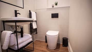 A compact bathroom with a white sink, black faucet, and a white toilet. There is a towel rack with folded towels, a small potted plant on the wall, and a black trash bin.