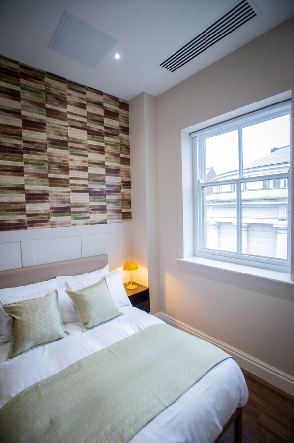 Un lit bien fait, recouvert d’une couvre-lit blanche et verte, avec une tête de lit et deux oreillers. Une fenêtre offre une vue sur les bâtiments à l’extérieur.